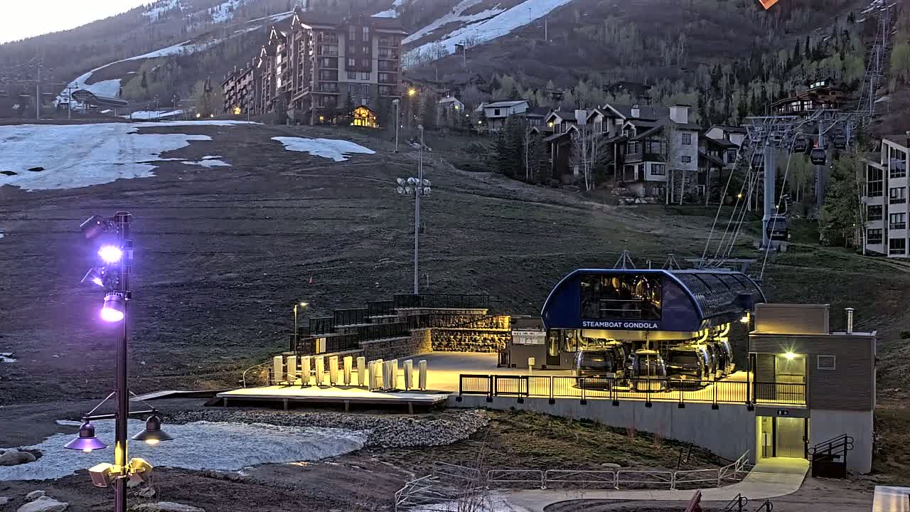 A gondola station at dusk sits below a ski resort with melting snow patches on the slopes and residential buildings on the hillside.