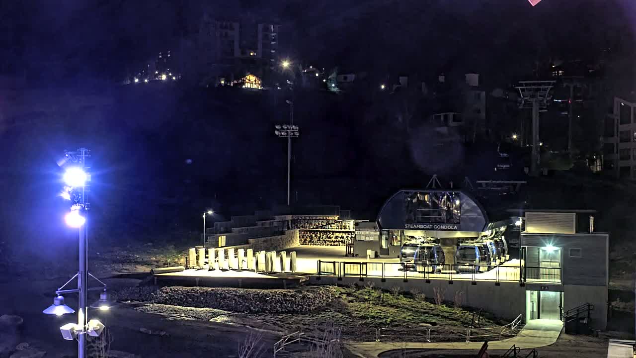 At night, a gondola station with several gondolas is illuminated, situated on a hillside with buildings and ski lifts in the background.