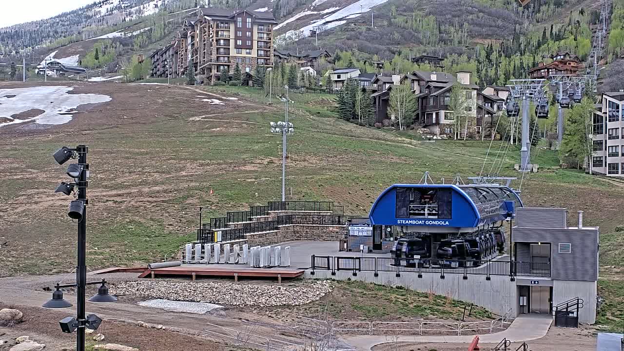 A gondola station sits at the base of a mountain with partially snow-covered slopes, numerous buildings, and some patchy green vegetation, under a mostly clear sky.