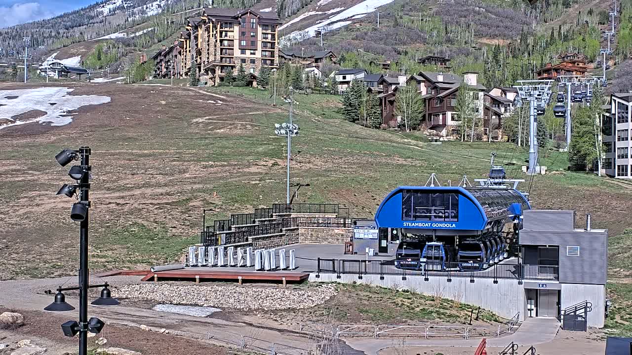 A blue gondola station sits at the base of a mountain on a sunny day, with patches of snow still visible on the hillside and surrounding buildings.