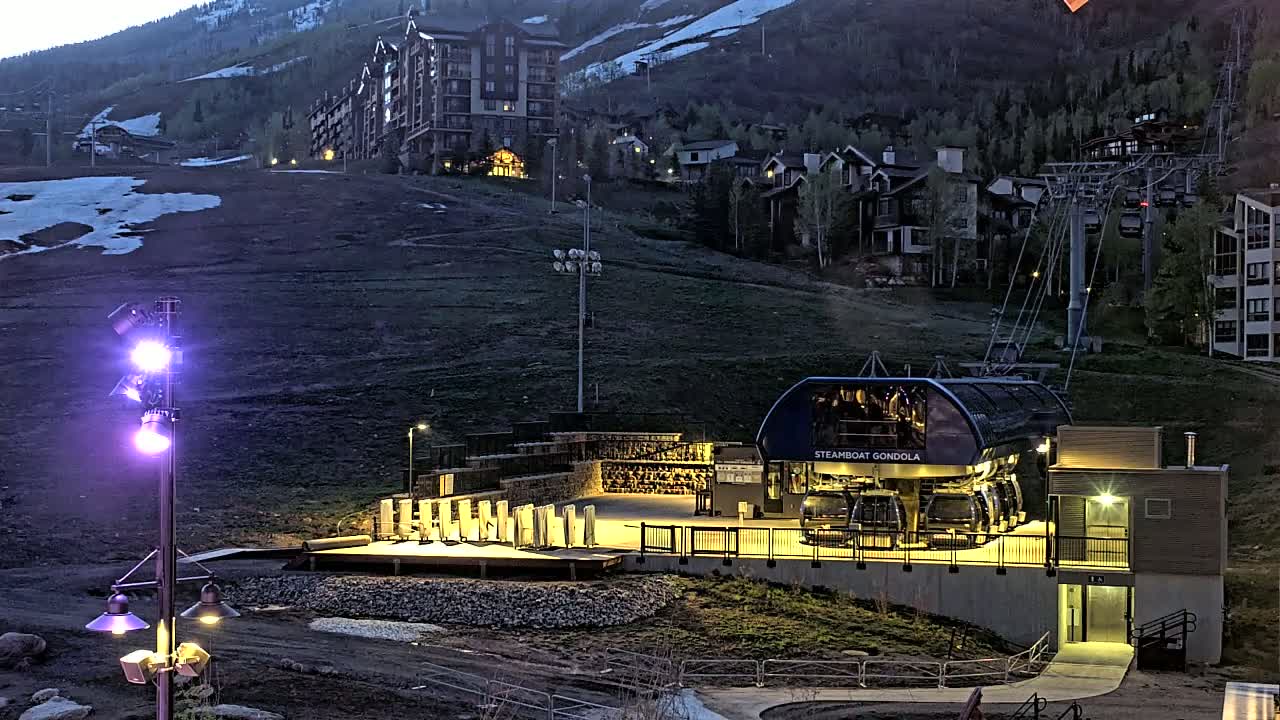 A gondola station at dusk, illuminated by artificial lights, sits below a residential area on a snow-dusted mountainside.