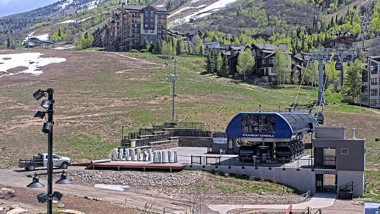 A gondola station sits below a hillside of green grass and buildings, with patches of snow visible on the mountainside in the background under a sunny sky.