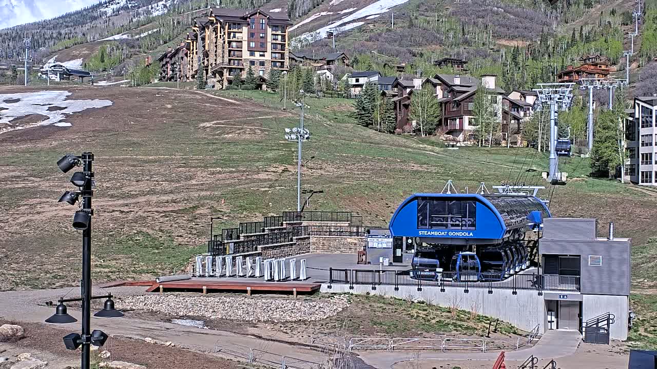 A blue gondola station sits below a hillside of residential buildings and patchy snow, under a sunny sky.