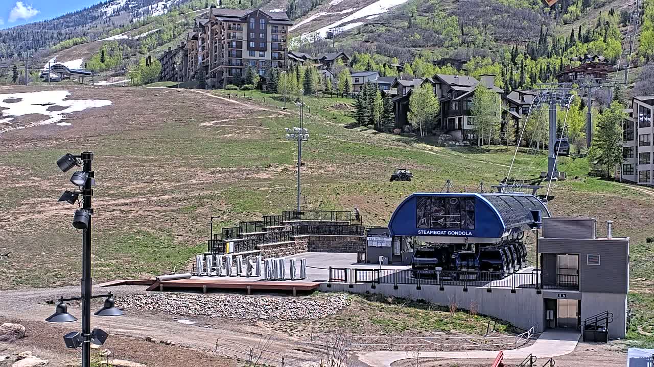 A gondola lift station sits below a hillside of green grass and buildings, with patches of snow visible on the mountainside in the background under a clear, sunny sky.