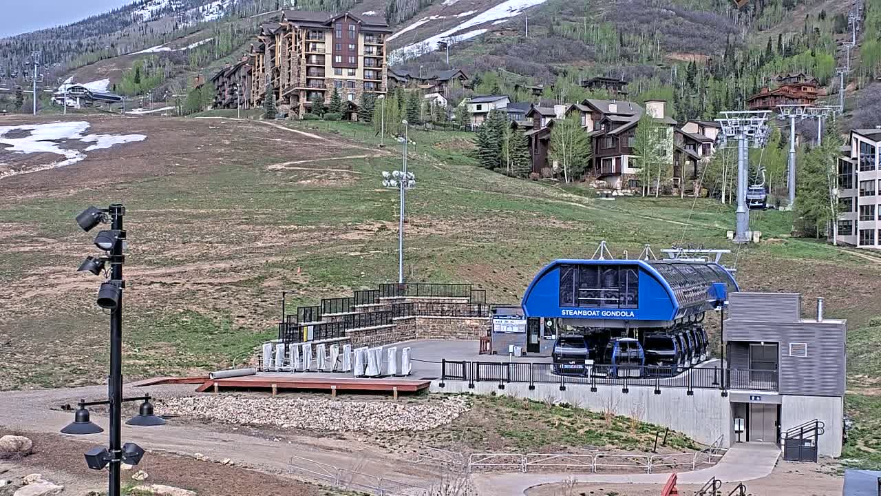 A gondola station sits below a hillside of residential buildings and ski lifts, with patchy snow visible on the mountainside in the background.