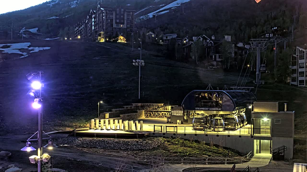 A gondola station, illuminated at night, sits below a slope with buildings and sparse snow, against a backdrop of dark mountains.