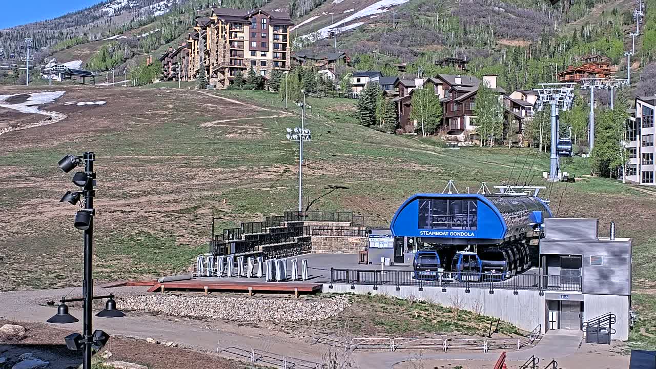A gondola station sits at the base of a mountain on a sunny day, with buildings and ski lifts visible in the background.