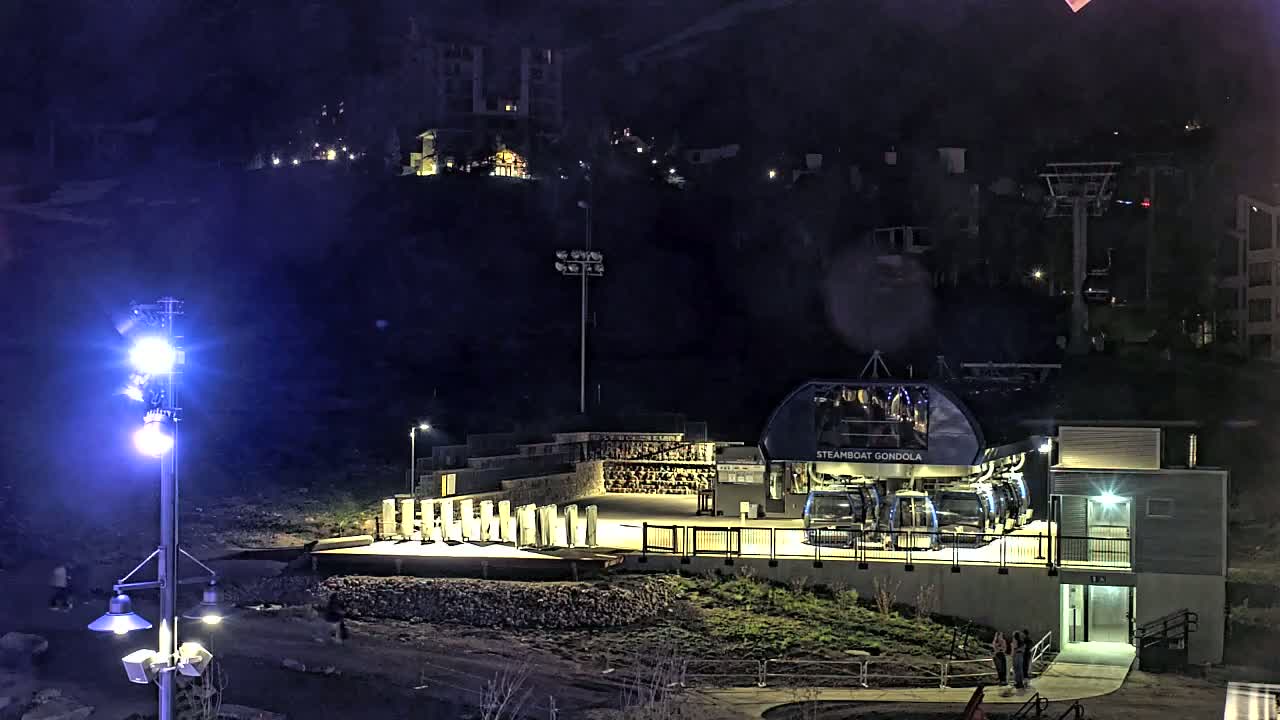 At night, a gondola station sits illuminated at the base of a hill with buildings and a ski lift visible in the distance.