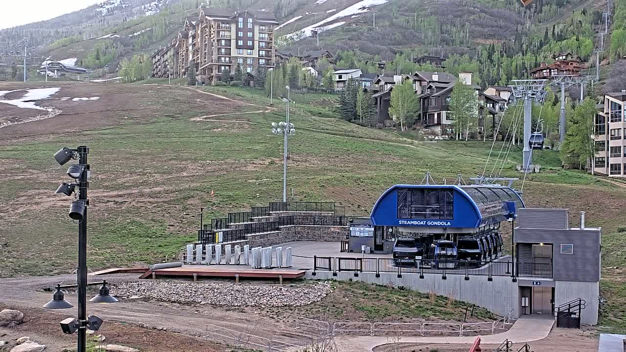 A gondola station sits on a grassy hillside overlooking a mountain village, with some snow still visible on the mountainside.