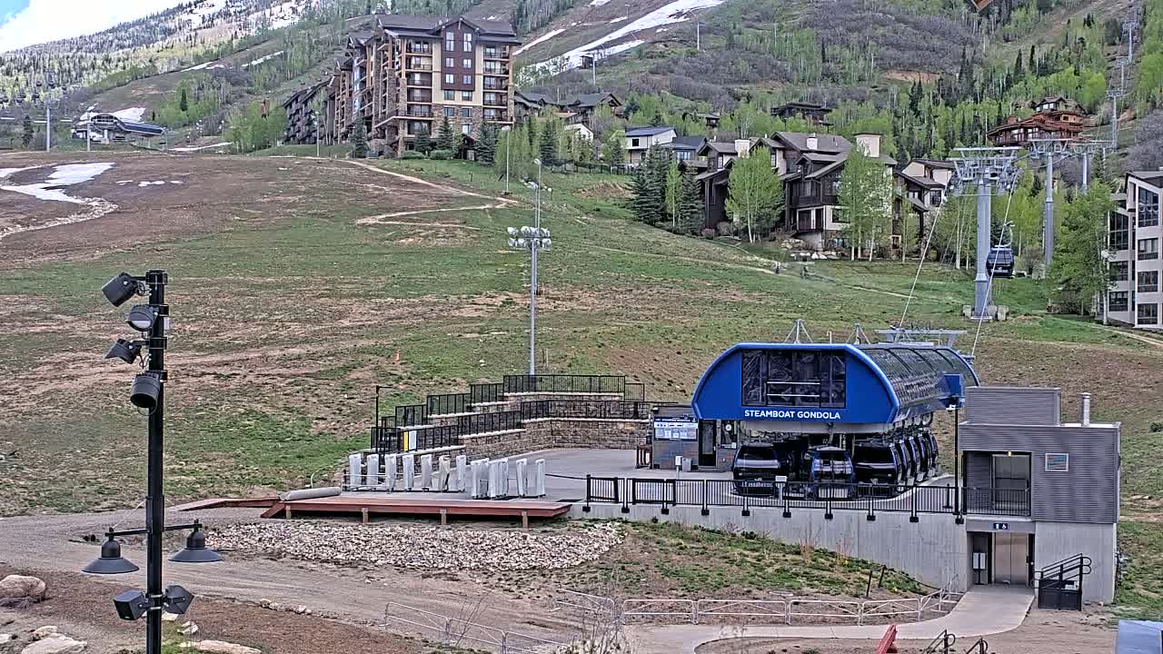 A gondola lift station sits below a hillside with residential buildings and a ski lift in the background on a partly sunny day with patches of snow remaining.