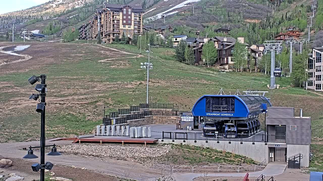A gondola station sits below a hill dotted with buildings and ski lifts, under a mostly clear sky.
