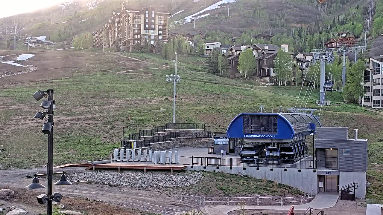 A gondola station sits on a grassy hillside beneath a mountain with buildings and a ski lift in the background, under mostly sunny conditions.