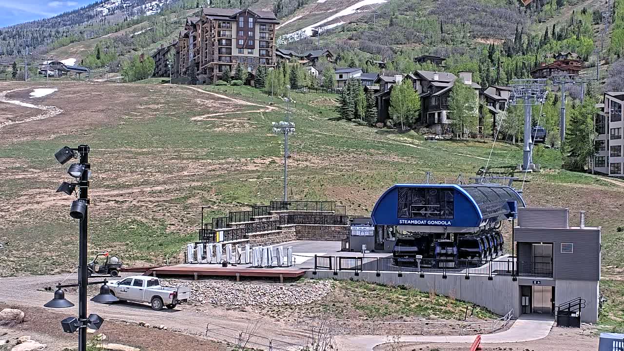 A gondola station sits below a hillside of buildings and grassy slopes on a sunny day.