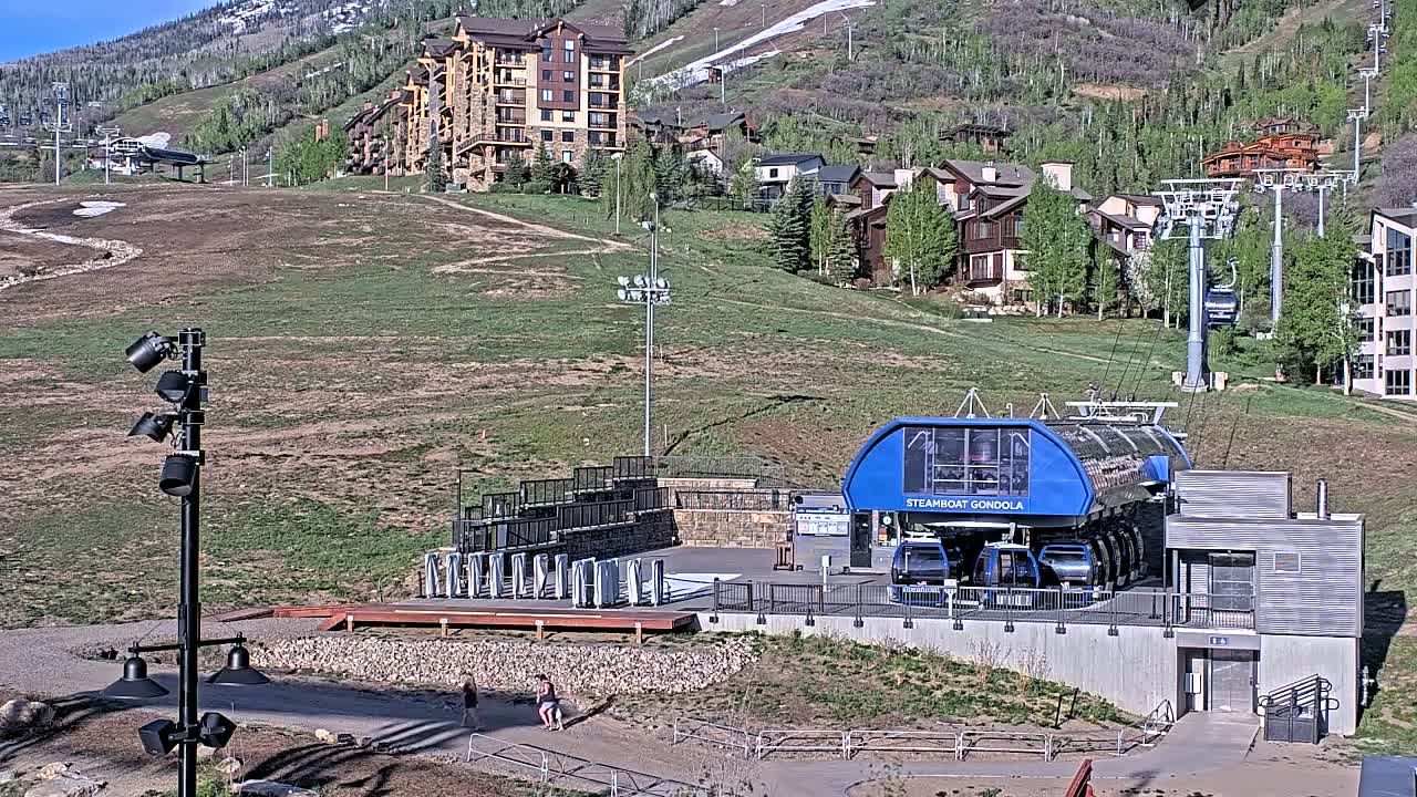 A gondola station sits below a hillside of buildings and ski lifts on a sunny day.