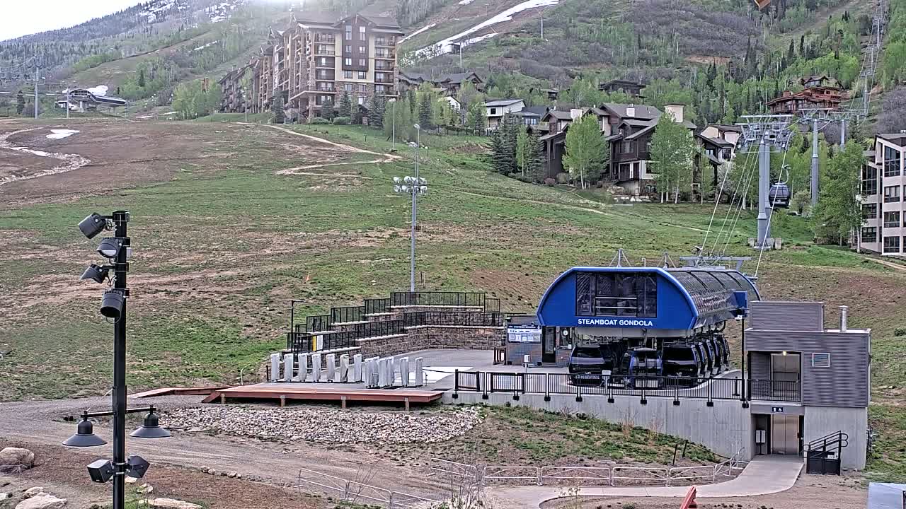 A gondola base station sits on a grassy hill overlooking a residential area, with a mountain in the background showing patches of snow under a mostly sunny sky.