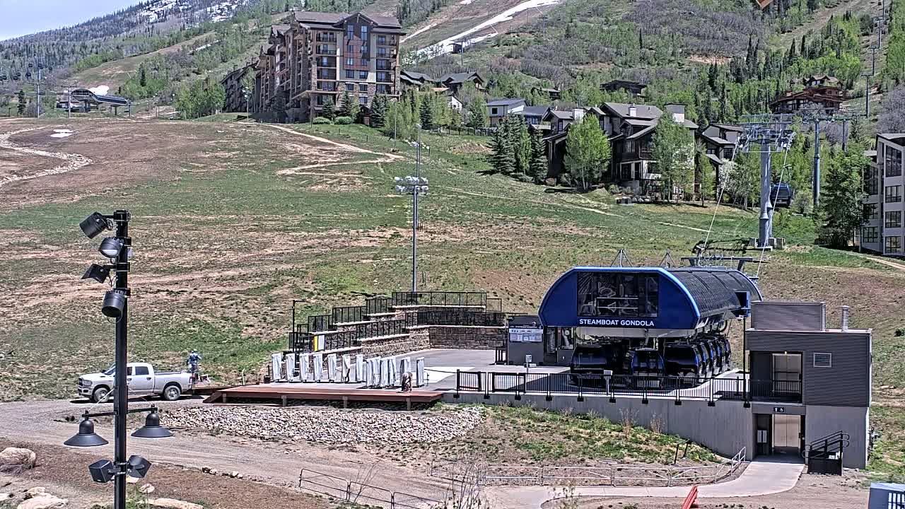 A gondola lift station sits below a residential area on a grassy hillside on a sunny day, with a truck parked nearby.