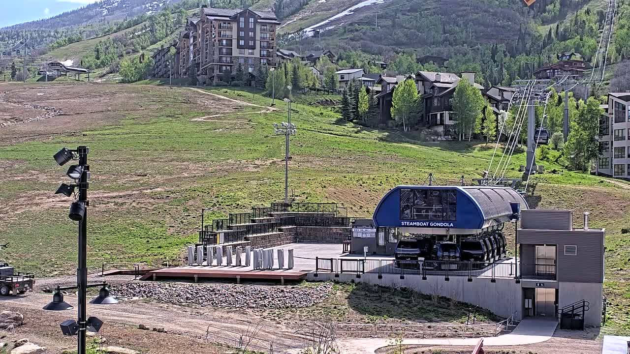 A gondola station sits at the base of a grassy mountainside dotted with buildings, under a mostly sunny sky.