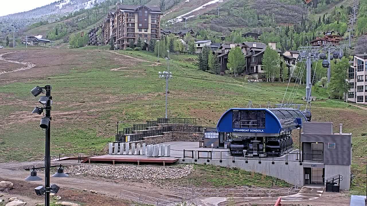 A gondola station sits below a hillside of residential buildings and a mountain partially covered in snow, under partly cloudy skies.