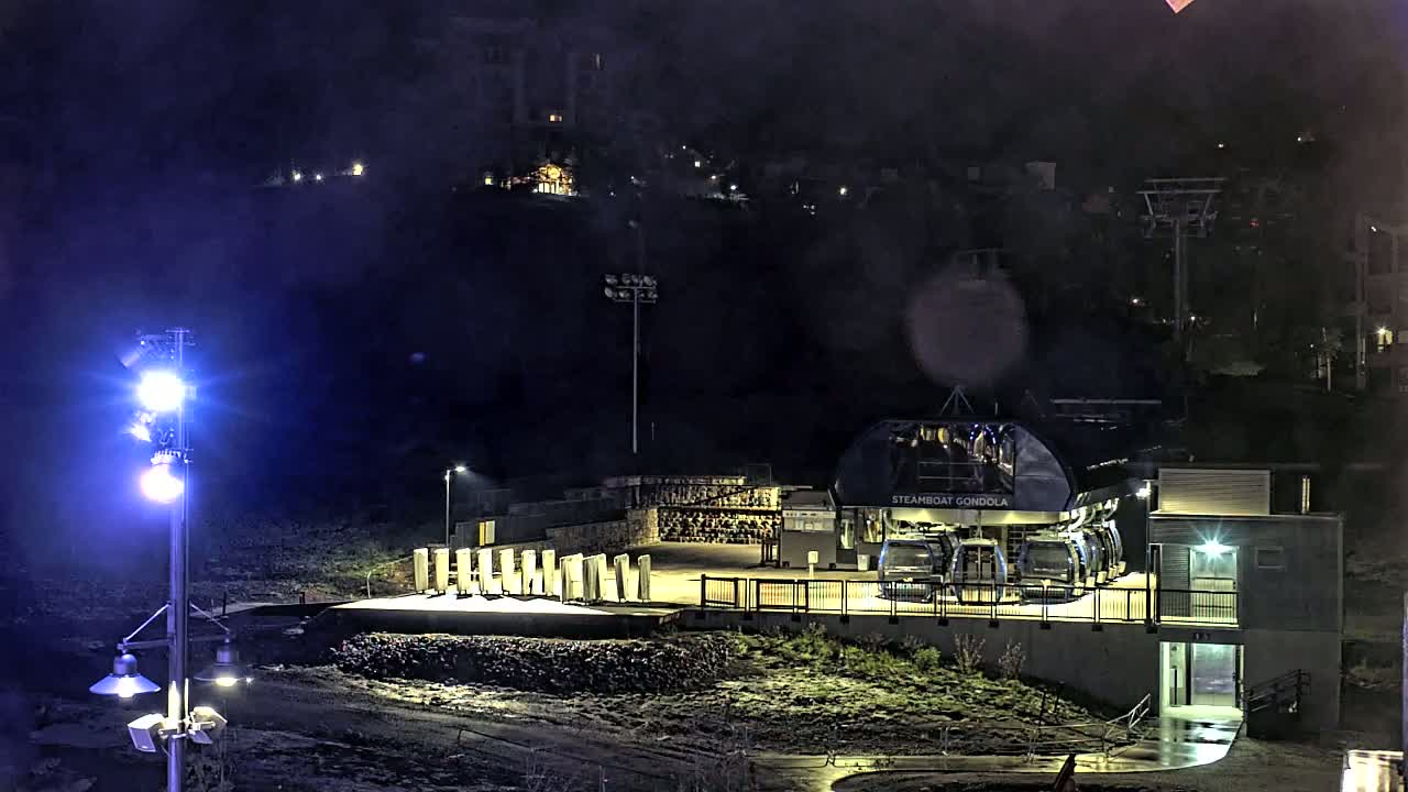 A gondola station is illuminated at night, with a nearby hilltop showing faint lights and a wet ground.