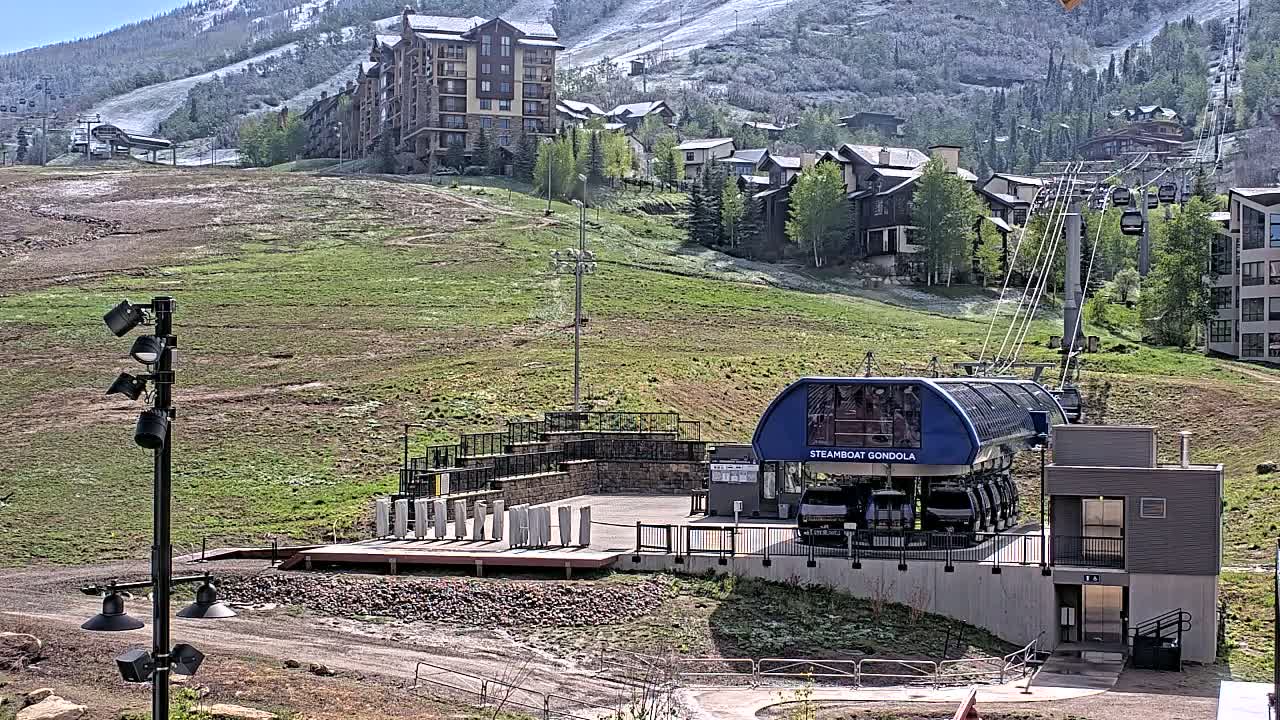 A gondola station sits on a grassy hillside below a partially snow-covered mountain, with buildings and a chairlift visible in the background under a sunny sky.