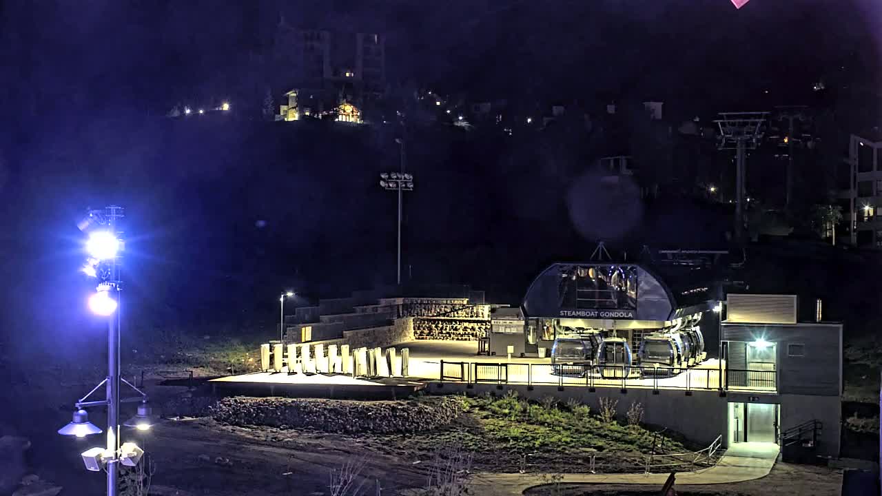 At night, a gondola station with illuminated cabins sits below a hillside of buildings, lit by bright lights under a dark sky.