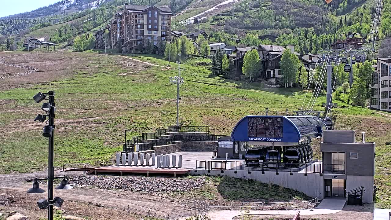 A gondola station sits at the base of a grassy hillside dotted with buildings, under a sunny sky.
