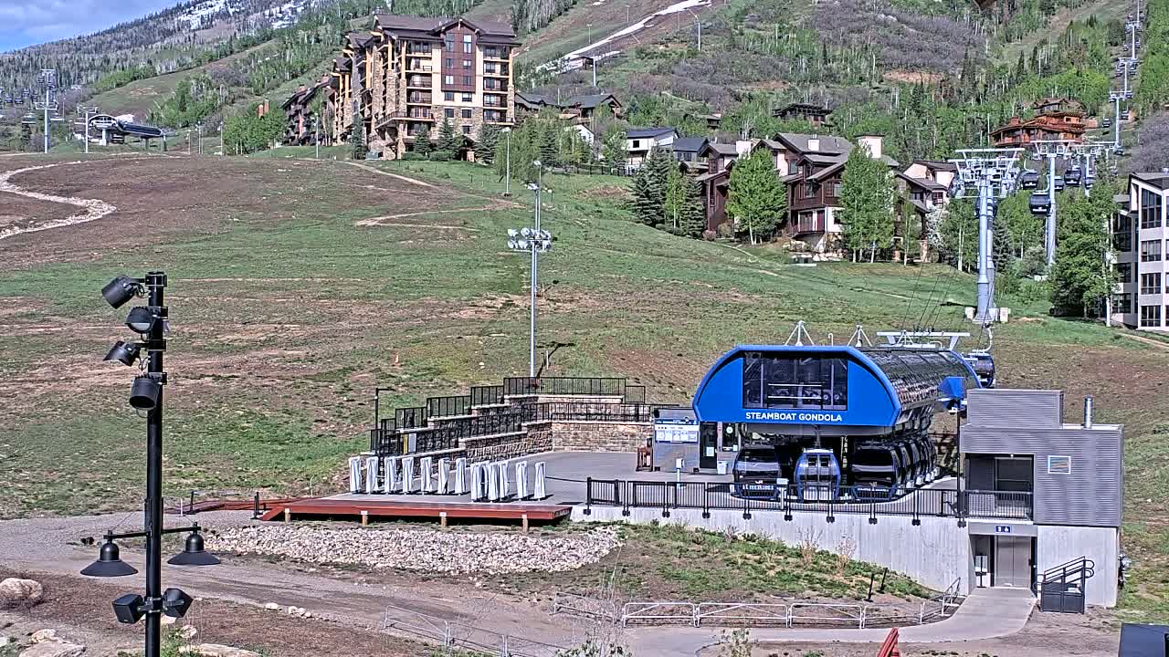 A blue gondola station sits below a hillside of houses and ski lifts on a sunny day with a mountain in the background.