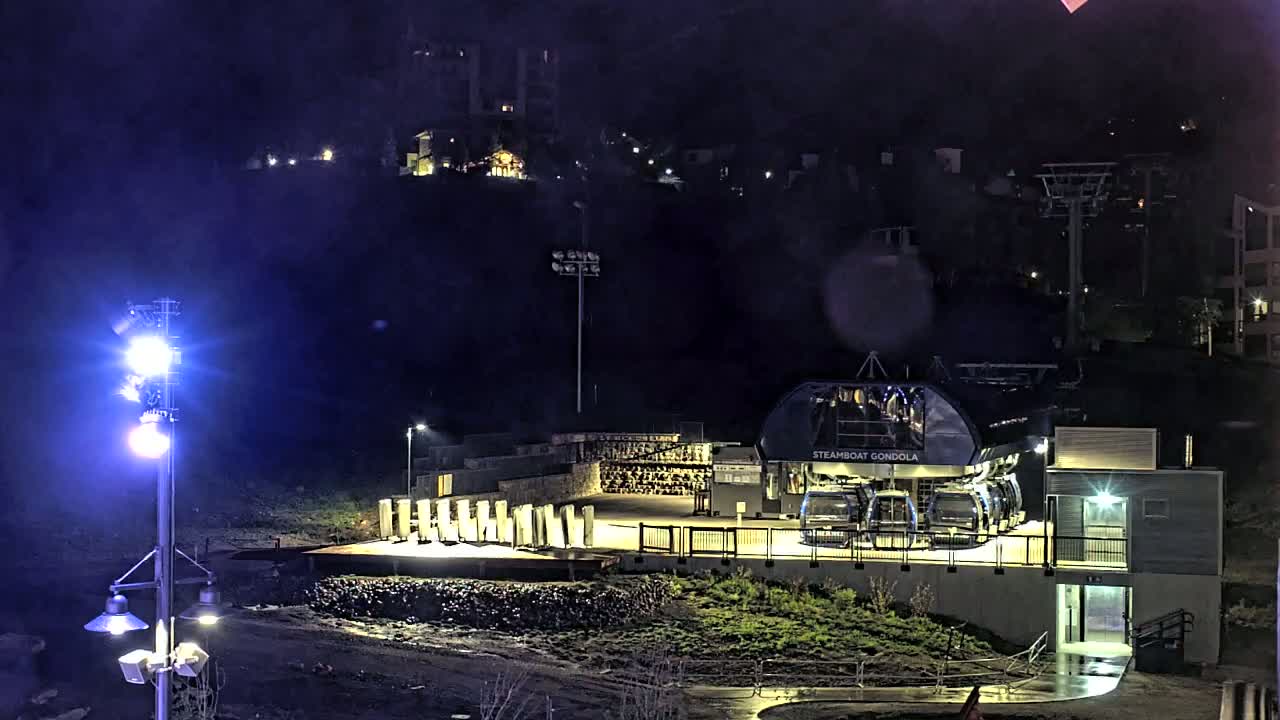 A gondola station at night is illuminated by bright lights, with rain or moisture visible on the ground, and a hillside with buildings in the background.