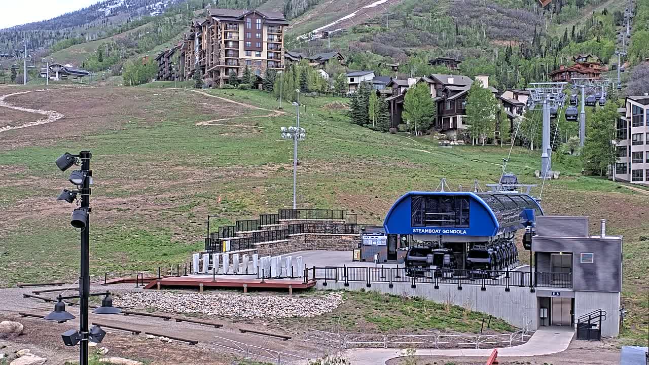 A gondola station sits at the base of a grassy hillside dotted with buildings, under a mostly clear sky.