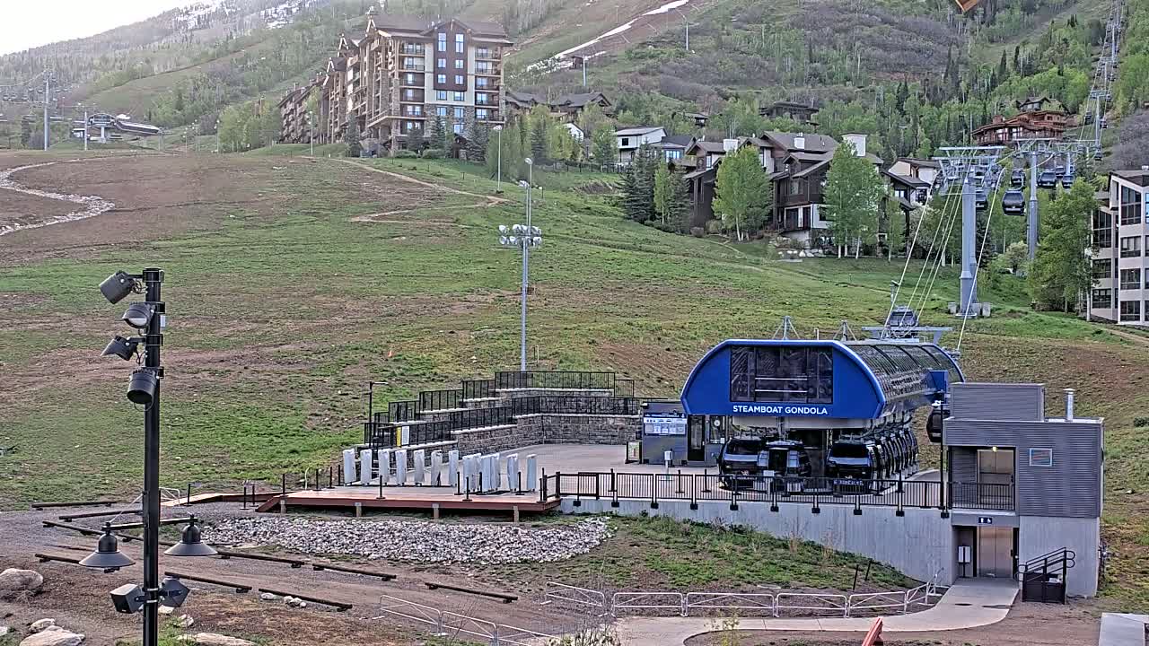 A gondola station sits on a grassy hillside, with residential buildings and a ski lift in the background, under a mostly clear sky.