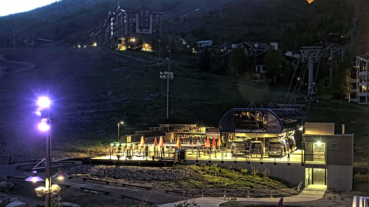 A gondola station at night, illuminated against a dark mountainside with several buildings visible in the background.