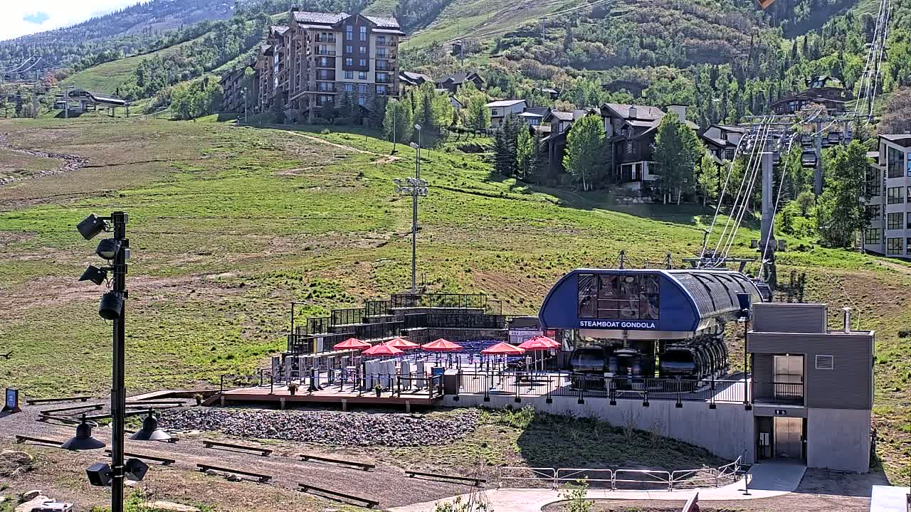 A gondola station with a dark-blue gondola car sits below a hillside of green grass and buildings, under a sunny sky.