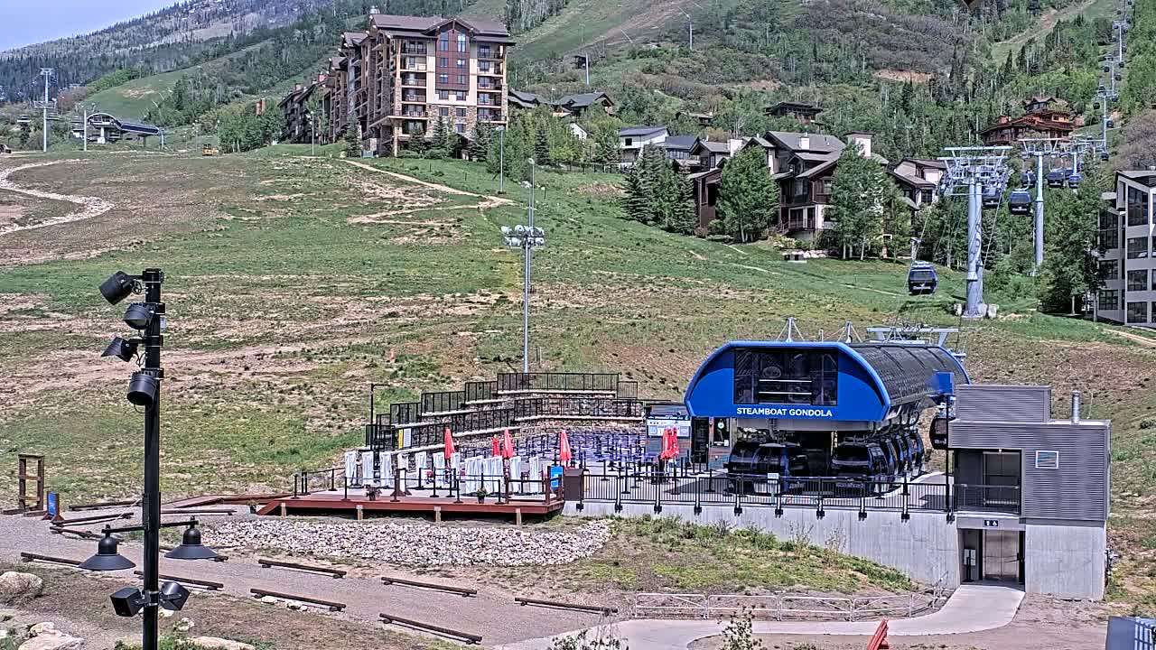 A gondola station sits below a residential area on a grassy hillside on a sunny day, with ski lifts visible in the background.