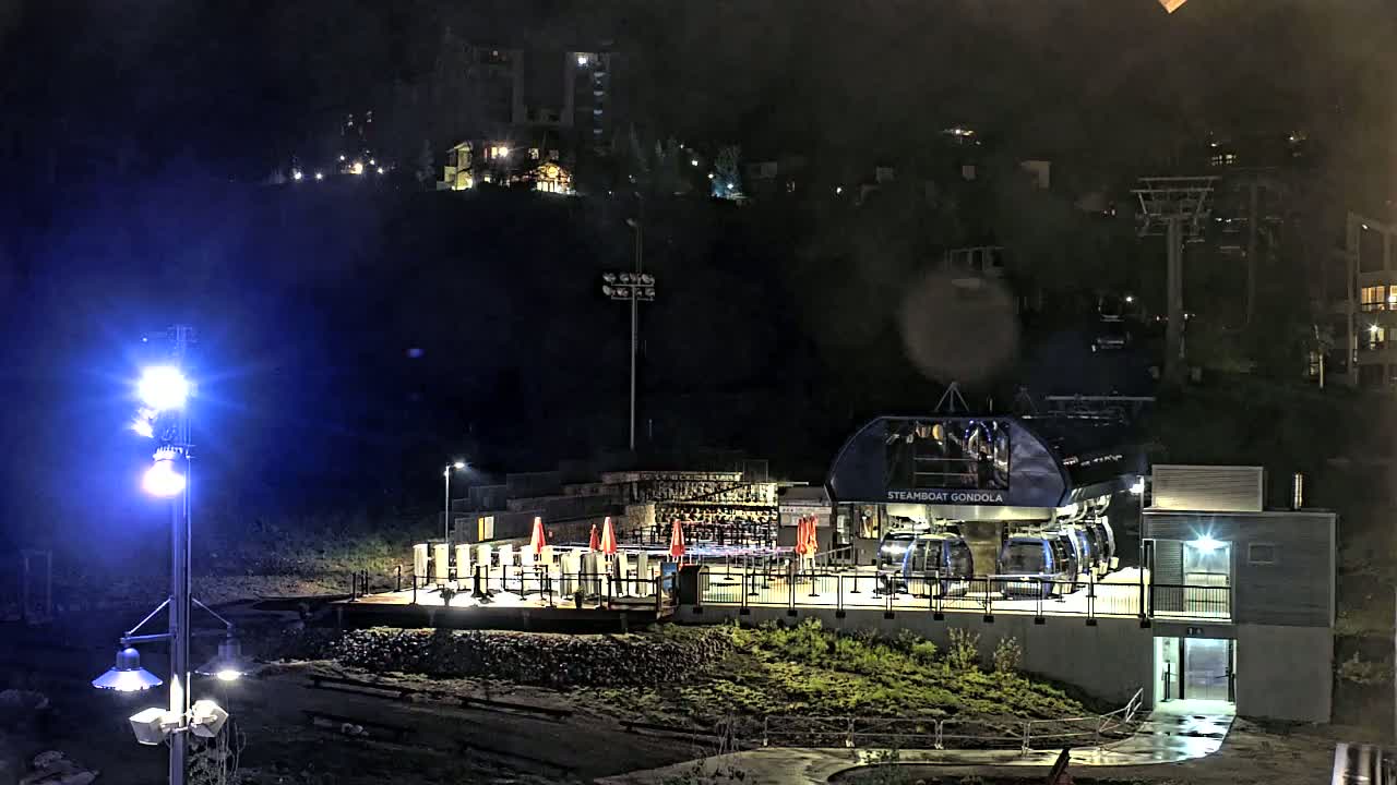 A gondola station is illuminated at night under rainy conditions, with a hillside of buildings visible in the background.