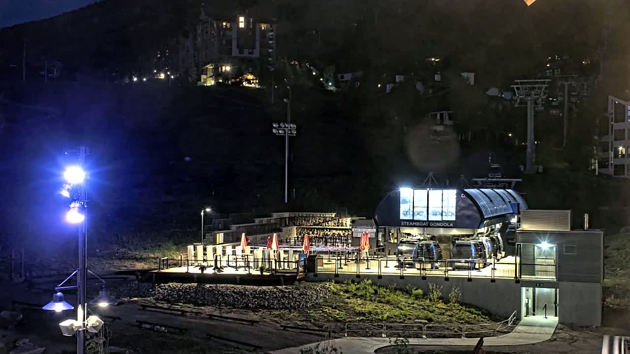 A gondola station at night, illuminated by bright lights, is situated on a hillside with buildings and ski lifts visible in the background.