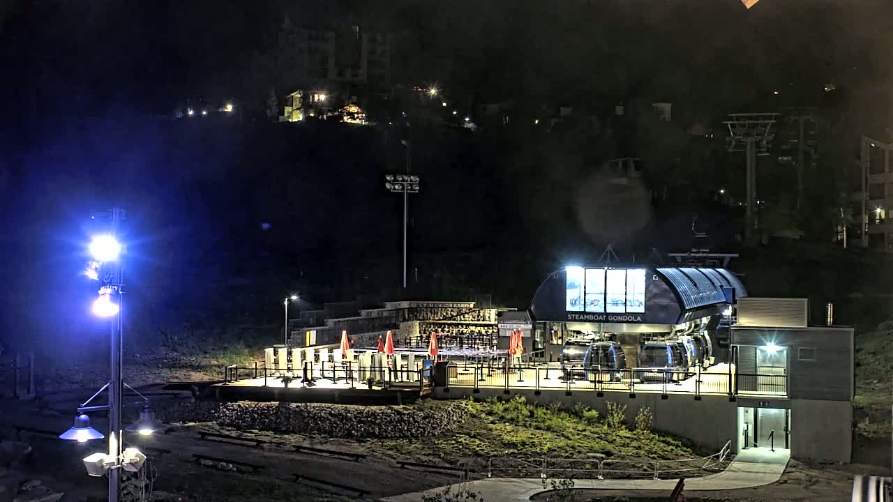 At night, a gondola station is brightly lit, situated on a hillside with buildings and a chairlift visible in the distance.
