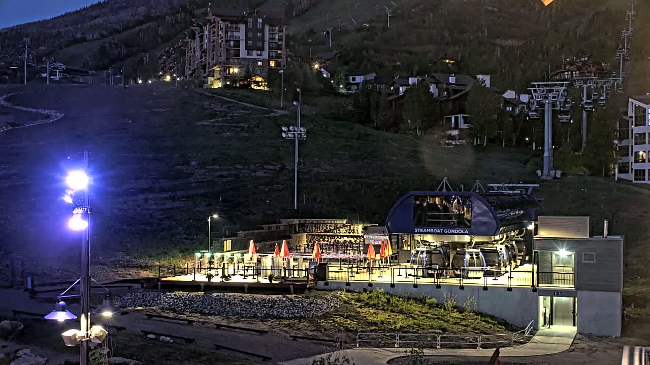 A gondola station at night, illuminated by artificial lights, sits on a hillside below buildings and ski lifts.