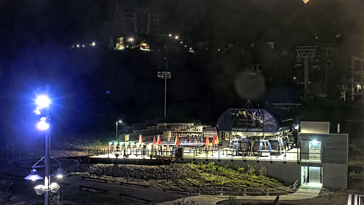 A gondola station is illuminated at night, with a hillside of buildings visible in the background under a dark, possibly rainy sky.