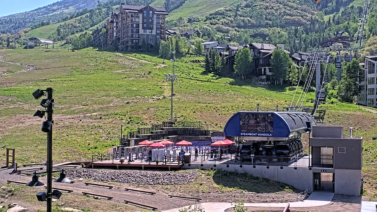 A gondola station sits on a grassy hillside below a cluster of buildings and a mountain range under a sunny sky.