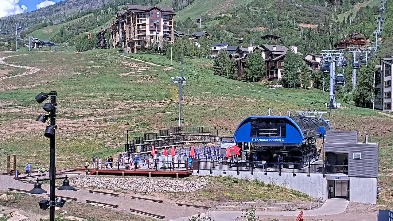 A gondola station with a blue gondola car sits at the base of a mountain on a sunny day, with several buildings and chairlifts visible in the background.