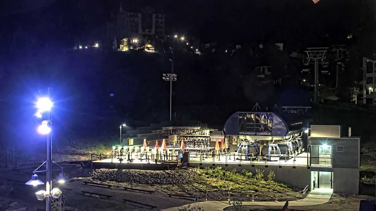 At night, a gondola station with its cabins illuminated is situated at the base of a hill with buildings visible on the hillside under a clear, dark sky.