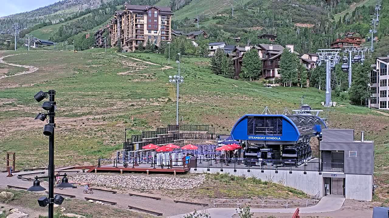A gondola station sits at the base of a mountain on a partly sunny day, with residential buildings and ski lifts visible on the hillside.