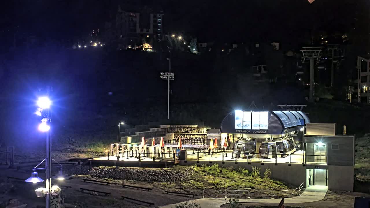 A gondola station, illuminated at night, sits at the base of a hill with buildings and ski lifts visible in the background.