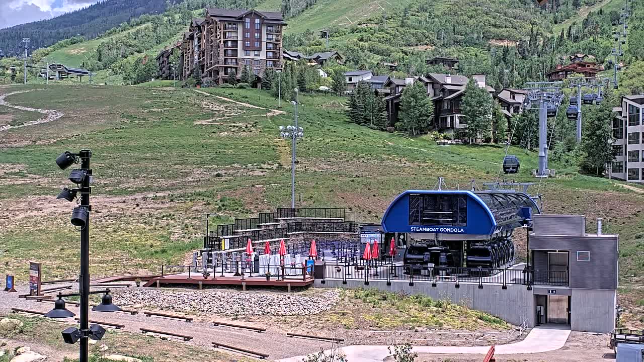 A gondola station sits on a grassy hillside beneath a cluster of buildings and a mountain, under a partly sunny sky.