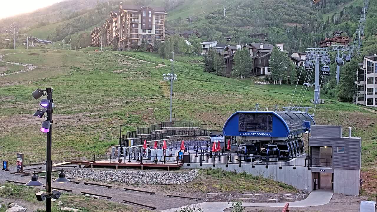 A gondola station sits at the base of a grassy hill dotted with buildings, under a clear evening sky.