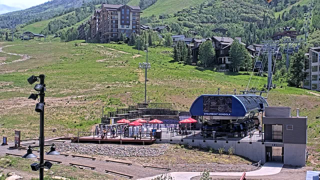 A gondola station with a blue gondola car sits below a grassy hillside dotted with buildings on a sunny day.