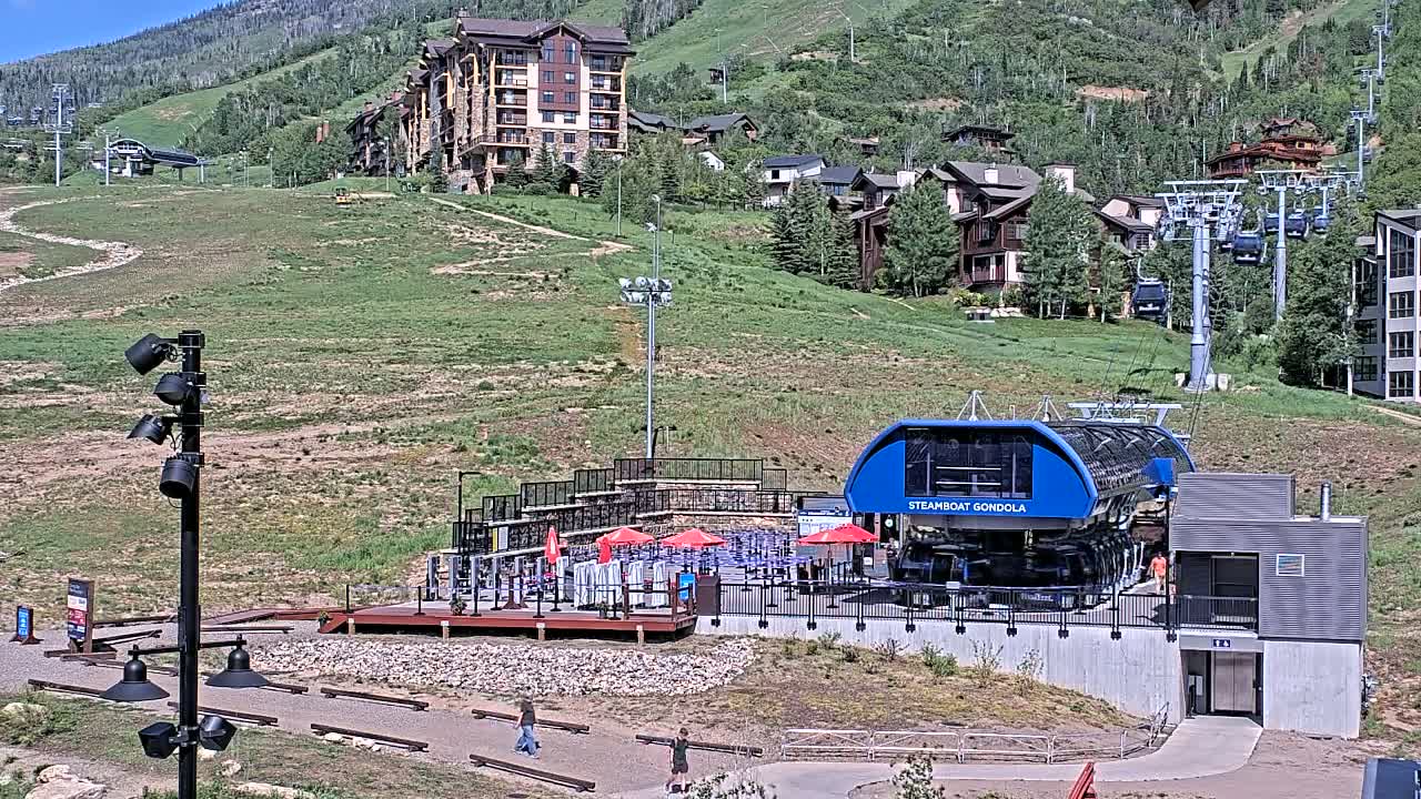 A blue gondola station sits at the base of a mountain on a sunny day, with several buildings and chairlifts visible in the background.