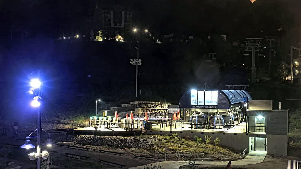 A gondola station illuminated at night, situated on a hillside with buildings and other lift infrastructure visible in the background.
