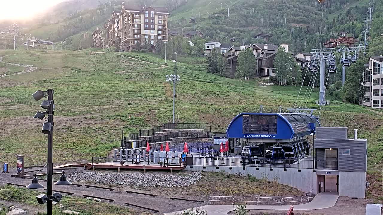 A gondola lift station sits at the base of a grassy hill dotted with buildings, under a partly sunny sky.