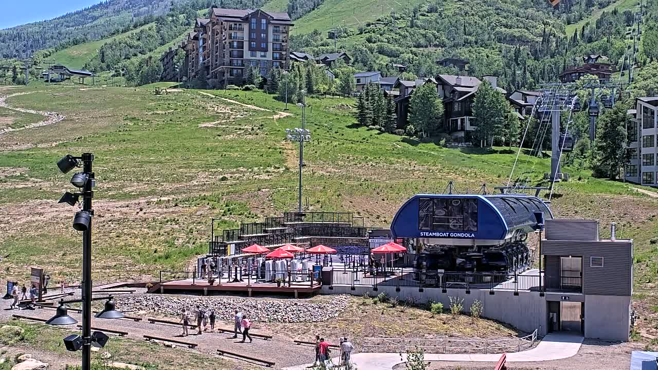 A gondola station with a blue gondola car sits on a grassy hillside beneath a sunny sky, with several buildings and a chairlift visible in the background.